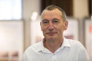 Edwin is a white man with short brown grey hair. He is wearing a white shirt and is smiling at the camera.